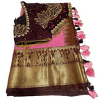 A folded traditional Indian ethnic wear fabric set featuring a deep maroon blouse with gold and silver zari embroidery and pearl accents, a bright pink inner fabric, and a wide gold brocade pallu with traditional motifs and pink tassels, displayed on a white marble-patterned surface.